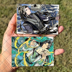 May include: Two rectangular enamel pins featuring anime-style characters. The top pin shows a figure with long black hair and dark clothing near a skeletal creature. The bottom pin depicts a character with long black hair holding a folding fan.