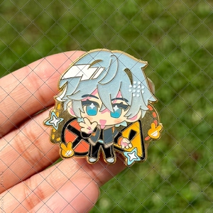 May include: A gold-toned enamel pin featuring a stylized anime-inspired character with light blue hair, blue eyes, and a dark jacket. The character is surrounded by decorative elements including a red circle with an X, a yellow triangle, and stars.