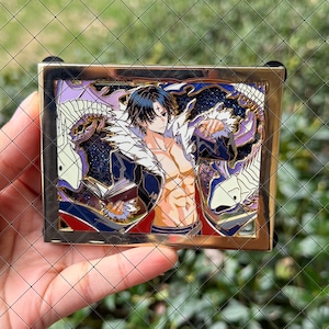 May include: A rectangular enamel pin featuring a dark-haired anime character with a cross on their forehead, shirtless, holding an open book. The character is surrounded by white dragon-like creatures against a dark blue glittery background.