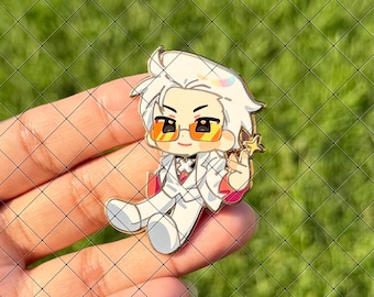 Peut inclure: Une broche en émail métallique représentant un personnage de style chibi aux cheveux blancs, portant des lunettes teintées orange et un costume blanc avec une cravate rouge. Le personnage tient une petite baguette étoile dorée.