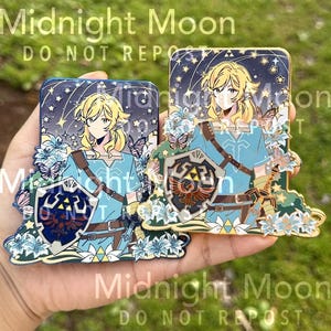 May include: Two enamel pins featuring a character with blonde hair, wearing a blue tunic, and holding a shield. The pins are decorated with flowers, butterflies, and a starry night sky. The text "Midnight Moon" and "DO NOT REPOST" are visible.