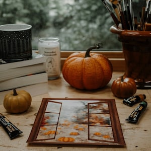 Peut inclure: Une nature morte automnale avec des citrouilles, des bougies, des livres et du matériel d'art. Une impression encadrée représente une fenêtre ouverte avec des citrouilles. Des tubes de peinture et des pinceaux sont également visibles.