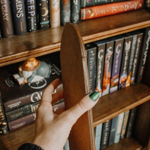 Op de afbeelding: Een houten boekenplank met verschillende boeken. Een hand houdt een bruine, gebogen houten plankverdeler vast. De boeken hebben titels als "SIREN", "WINTERWOOD" en "DECK OF OMENS". De verdeler is gemaakt van gelaagd hout.