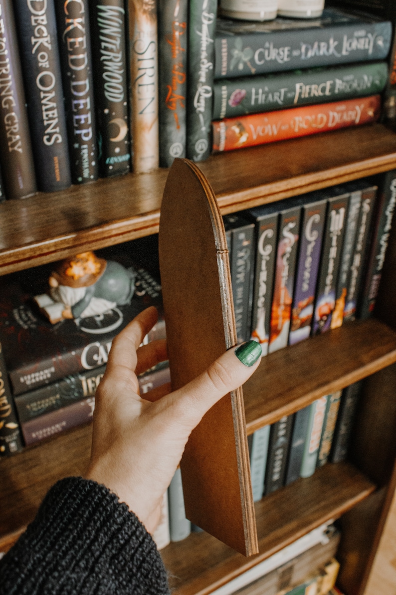 Op de afbeelding: Een houten boekenplank met verschillende boeken. Een hand houdt een bruine, gebogen houten plankverdeler vast. De boeken hebben titels als "SIREN", "WINTERWOOD" en "DECK OF OMENS". De verdeler is gemaakt van gelaagd hout.