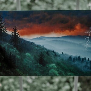 以下が含まれることがあります： ドラマチックな空の下の山脈を描いた風景画。鮮やかなオレンジと赤の夕焼けと、シーンを照らす稲妻が特徴です。前景には濃い緑色の木々が描かれています。