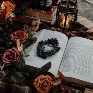 May include: An open book with a black ornate frame with a moth illustration inside. The book is on a plaid blanket with a lantern and fall-themed flowers.