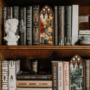 Op de afbeelding: Een donkerhouten boekenkast met een verzameling hardcover boeken. Titels als "Ninth House" en "Crooked Kingdom" zijn zichtbaar. Decoratieve spiegels en een klassieke buste dragen bij aan de esthetiek. Een kaars met het woord "Present" is ook aanwezig.