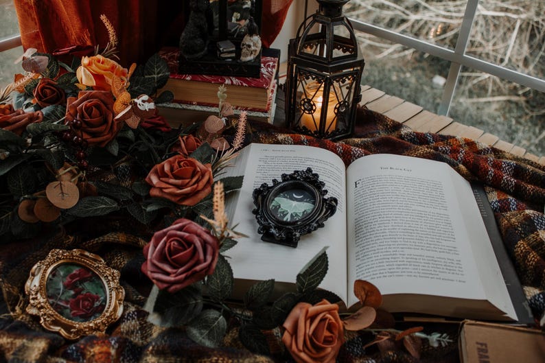 May include: An open book with a black framed picture of a moth on the cover. The book is surrounded by a bouquet of red and orange roses, a small gold picture frame, and a black lantern.