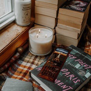 May include: A book titled "A Paris Apartment" by Lucy Foley, a New York Times Bestseller, with a bookmark featuring a photo of the Eiffel Tower. The book is on a plaid blanket with two candles, one labeled "November Seventeen" and the other with three wicks.