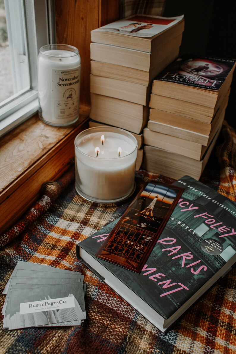 May include: A book titled "A Paris Apartment" by Lucy Foley, a New York Times Bestseller, with a bookmark featuring a photo of the Eiffel Tower. The book is on a plaid blanket with two candles, one labeled "November Seventeen" and the other with three wicks.