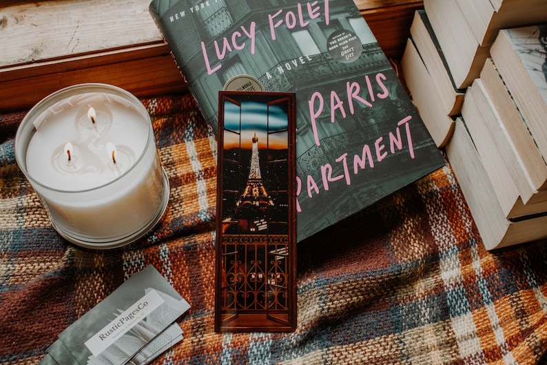 May include: A book titled "Lucy Foley: Paris Apartment" with a bookmark featuring a view of the Eiffel Tower at night. The book and bookmark are on a plaid blanket with a candle.