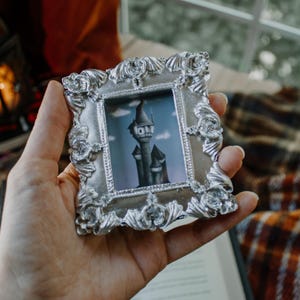 May include: A small, ornate silver picture frame with a detailed border. The frame holds a miniature painting of a castle with a blue sky background. The frame is held in a hand, suggesting a decorative item or keepsake.