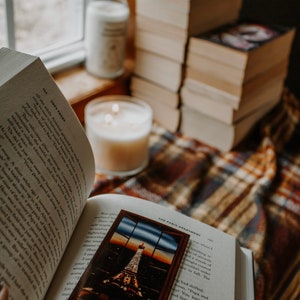 May include: An open book with a bookmark featuring a view of the Eiffel Tower in Paris, France. The book is resting on a plaid blanket with two candles nearby.