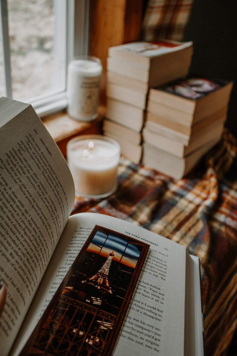 May include: An open book with a bookmark featuring a view of the Eiffel Tower in Paris, France. The book is resting on a plaid blanket with two candles nearby.
