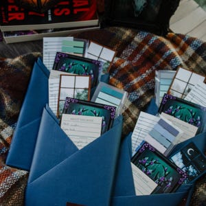 Peut inclure: Des enveloppes en papier bleu foncé remplies de cartes et de notes, certaines avec des illustrations d'une scène de forêt. Une image encadrée et un livre sont visibles en arrière-plan. Les enveloppes ont un autocollant Cryptids.