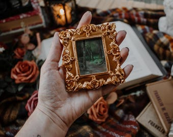 May include: A small, ornate gold-colored picture frame with a miniature landscape painting. The frame has a detailed floral design. The image is held in a hand, with roses and books in the background.