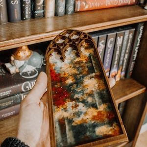 Op de afbeelding: Een houten kunstwerk met een kleurrijk abstract ontwerp. Het kunstwerk heeft een mix van rode, oranje en witte tinten, die lijken op een landschap. Het frame heeft een gewelfde bovenkant. Boekenplanken zijn op de achtergrond.