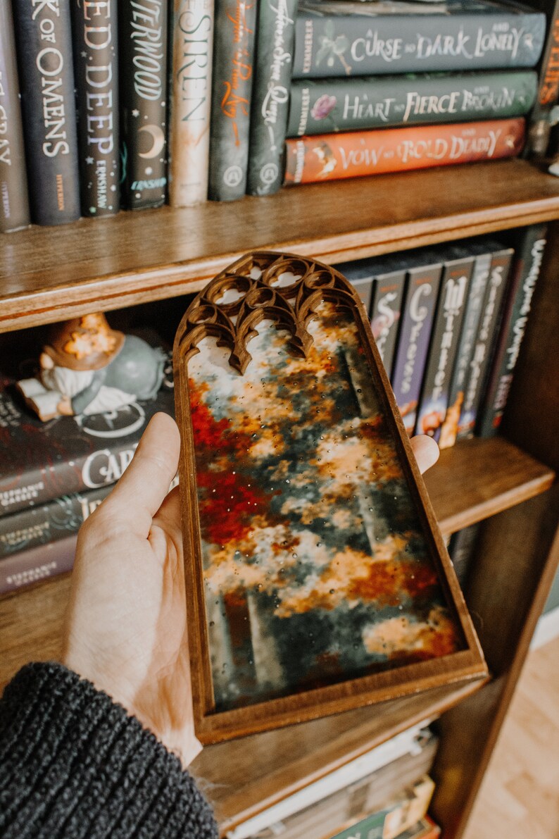 Op de afbeelding: Een houten kunstwerk met een kleurrijk abstract ontwerp. Het kunstwerk heeft een mix van rode, oranje en witte tinten, die lijken op een landschap. Het frame heeft een gewelfde bovenkant. Boekenplanken zijn op de achtergrond.