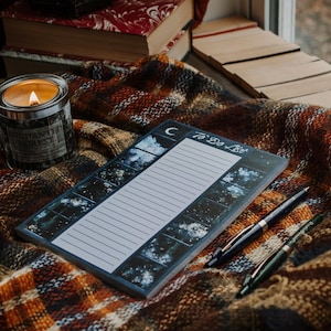 May include: A black and white to-do list notepad with a crescent moon and stars design. The notepad is lying on a plaid blanket with two pens next to it.