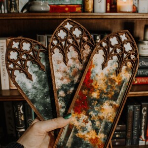 Op de afbeelding: Drie houten bladwijzers met een glas-in-lood ontwerp worden vastgehouden. De bladwijzers hebben een donkerhouten frame en ingewikkelde uitgesneden patronen. Het glas-in-lood heeft een mix van kleuren, waaronder groen, rood en geel, met een wolkeneffect.