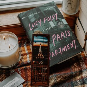 May include: A book titled "Paris Apartment" by Lucy Foley, a New York Times bestselling author, with a bookmark featuring the Eiffel Tower at sunset. The book is on a plaid blanket with a candle and a stack of books.