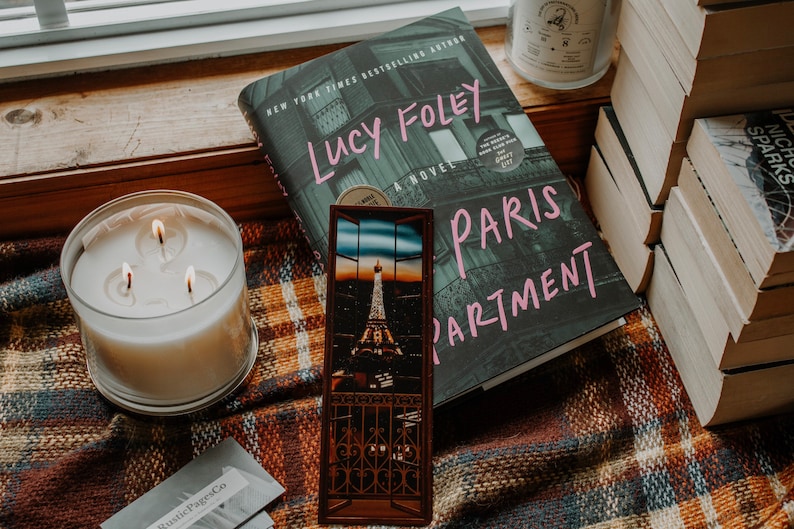 May include: A book titled "Paris Apartment" by Lucy Foley, a New York Times bestselling author, with a bookmark featuring the Eiffel Tower at sunset. The book is on a plaid blanket with a candle and a stack of books.