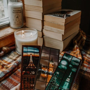 May include: Three bookmarks with cityscapes. The bookmarks are made of a dark brown material with a clear plastic window. The first bookmark shows the Eiffel Tower in Paris, the second bookmark shows Big Ben in London, and the third bookmark shows the Empire State Building in New York City.