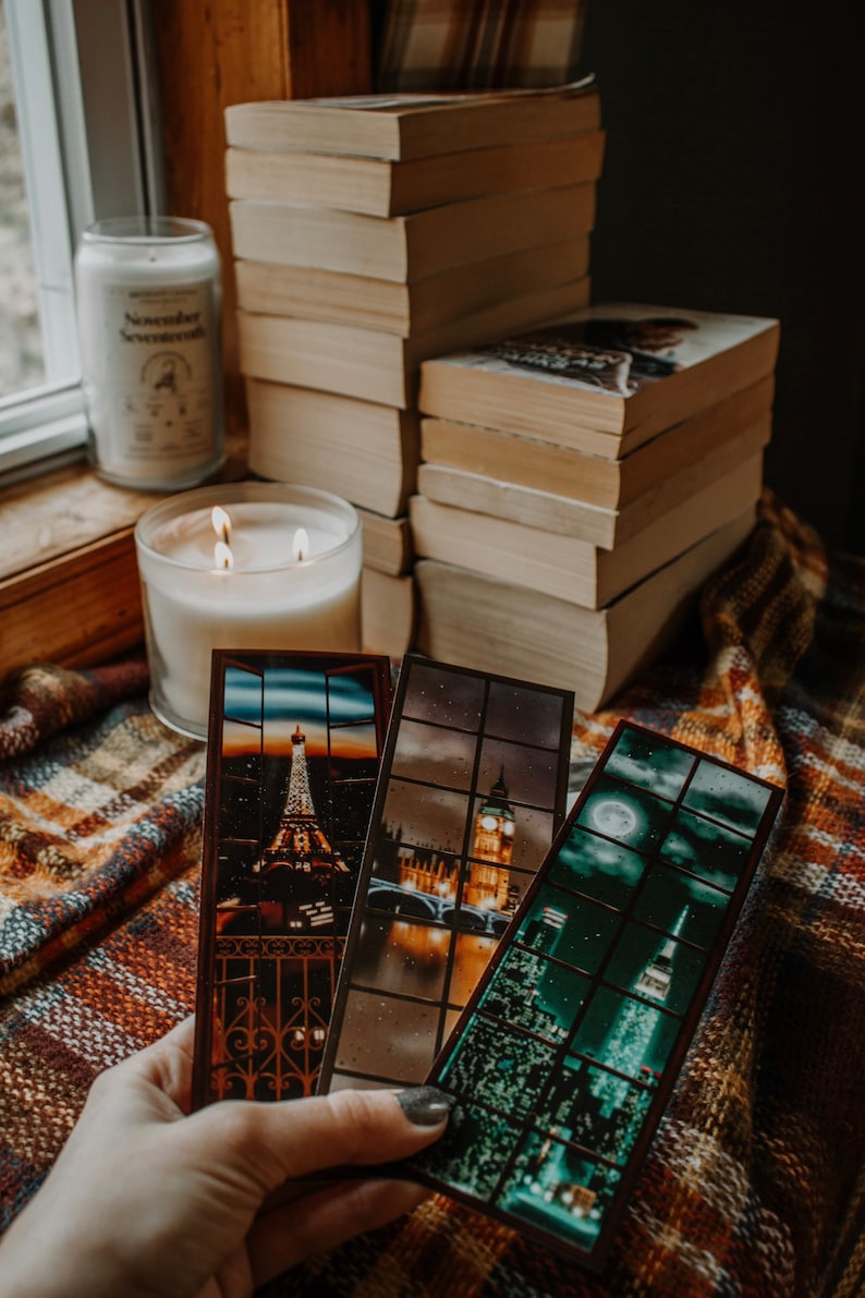 May include: Three bookmarks with cityscapes. The bookmarks are made of a dark brown material with a clear plastic window. The first bookmark shows the Eiffel Tower in Paris, the second bookmark shows Big Ben in London, and the third bookmark shows the Empire State Building in New York City.