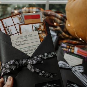 May include: Black envelope-style gift box with a white spiderweb ribbon. The box contains a "Young Adult Paperback" book and other items. A "Horror" label is on the front. Another box with a "Mystery/Thriller" label is also visible.