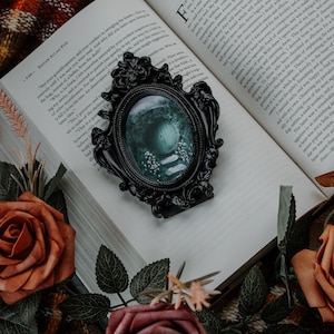 May include: A black ornate picture frame with a green and white image inside. The frame is sitting on top of a book with a plaid blanket and flowers in the background.