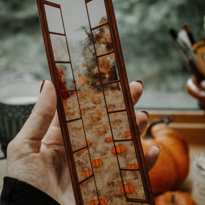 May include: A brown wooden bookmark with a window-like design. The window is open and reveals a field of orange pumpkins in a blurry background.