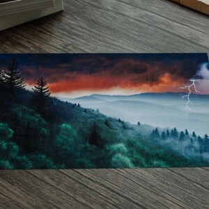 以下が含まれることがあります： 劇的な空の下の山脈を描いた風景画。このアートワークは、霧のかかった森林の丘の上に稲妻が走る鮮やかな夕焼けを特徴としています。カラーパレットには、緑、青、オレンジの色合いが含まれています。