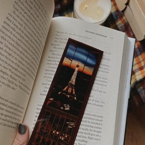 May include: A brown wooden bookmark with a picture of the Eiffel Tower at sunset. The bookmark is in front of an open book with the text "The Paris Apartment" on the page.