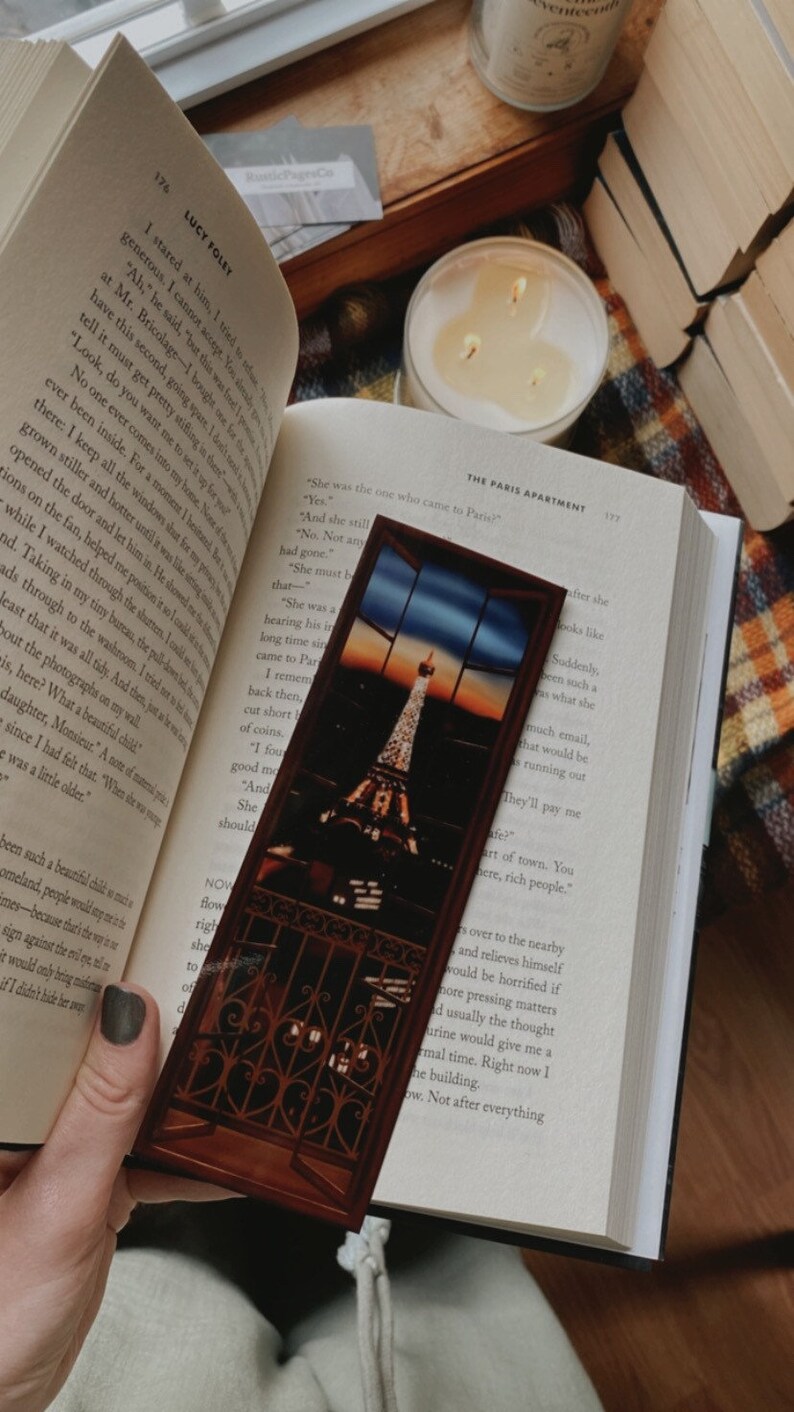 May include: A brown wooden bookmark with a picture of the Eiffel Tower at sunset. The bookmark is in front of an open book with the text "The Paris Apartment" on the page.