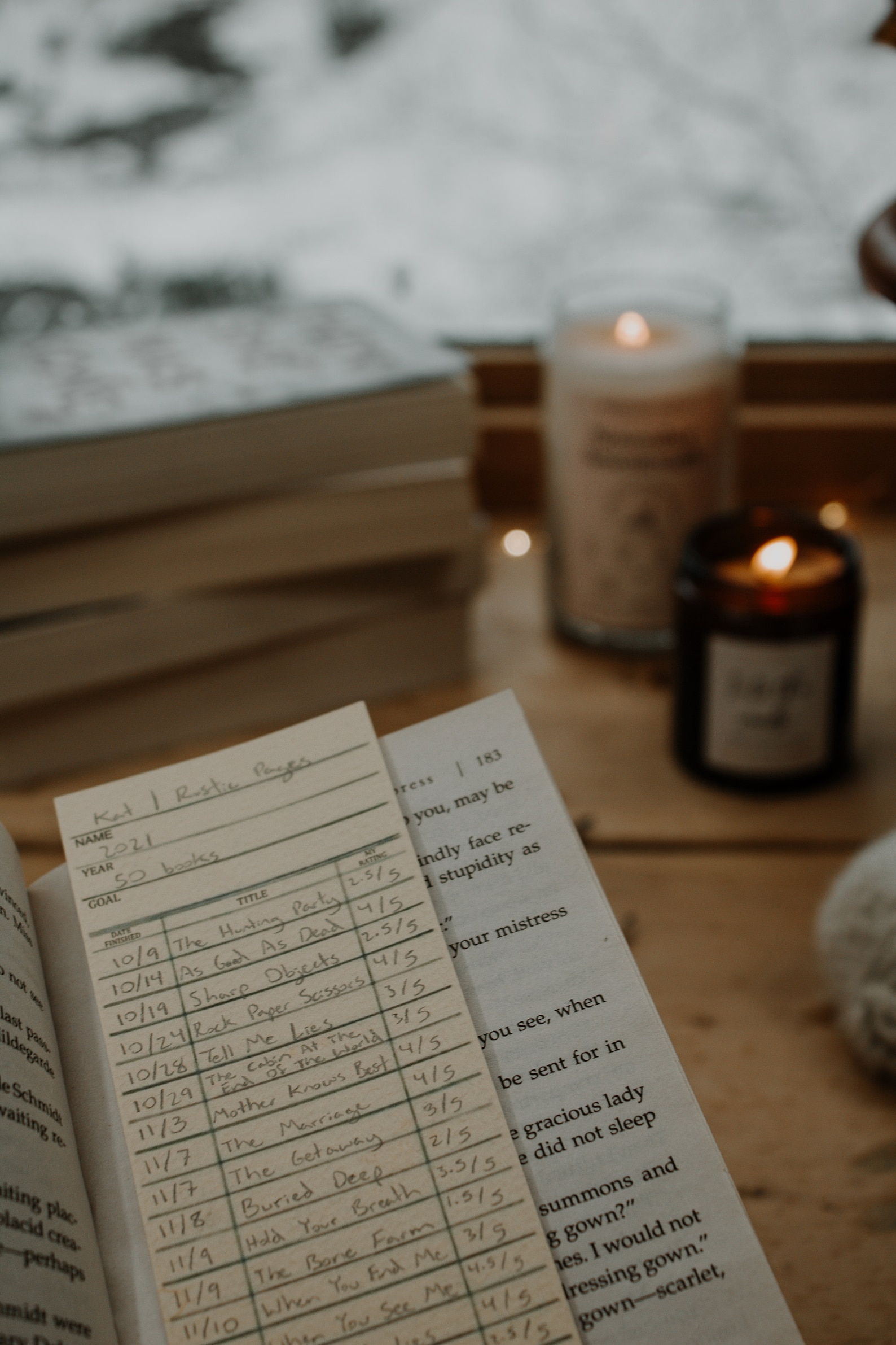 Open book with reading list sits on a wooden table. Two lit candles add warmth. Stacked books in the background by a snowy window create a cozy ambiance.