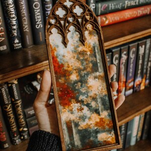 Op de afbeelding: Een houten kunstwerk met een glas-in-lood effect. Het kunstwerk toont een bewolkt landschap met tinten oranje, rood en grijs. Het frame heeft een gewelfde bovenkant met decoratieve uitsparingen.