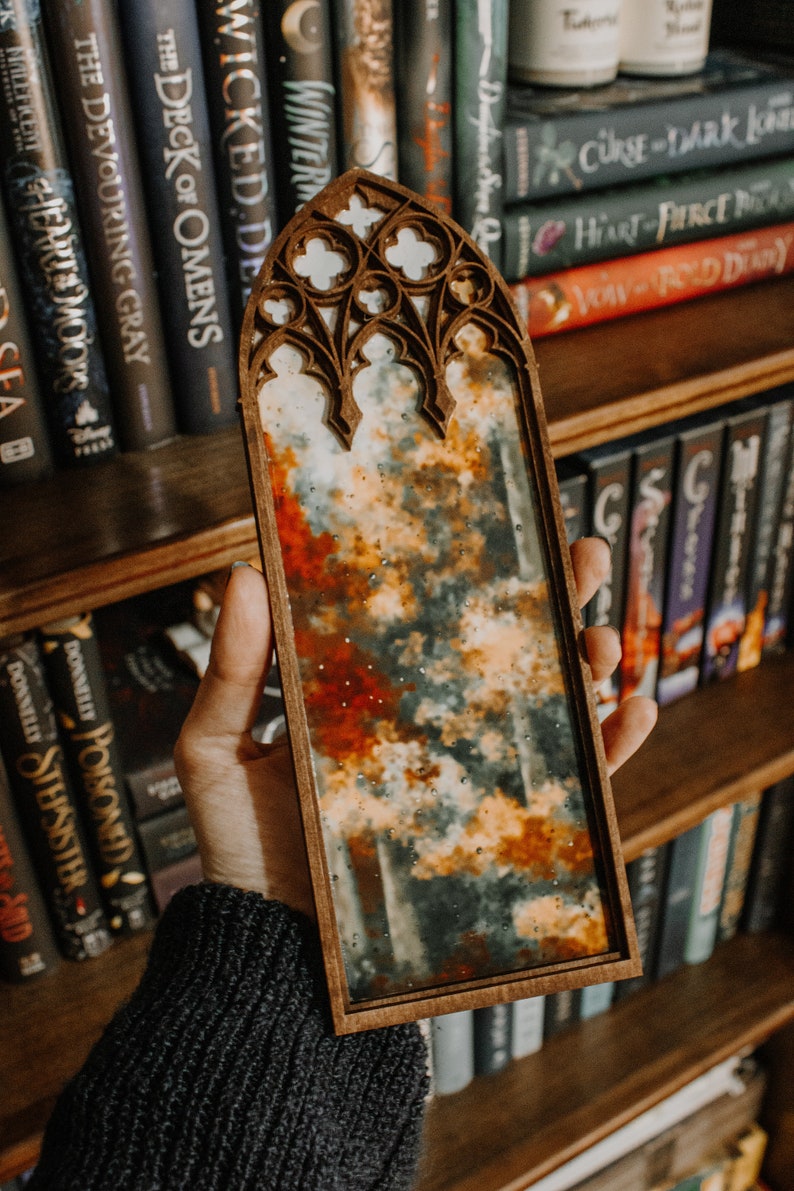 Op de afbeelding: Een houten kunstwerk met een glas-in-lood effect. Het kunstwerk toont een bewolkt landschap met tinten oranje, rood en grijs. Het frame heeft een gewelfde bovenkant met decoratieve uitsparingen.