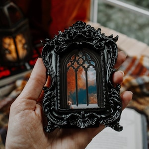 May include: A miniature black framed picture with a gothic window design. The window shows a colourful sky with orange and blue tones. The frame is held in a hand, suggesting a small decorative item or artwork.
