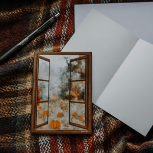 May include: A greeting card with a watercolor painting of pumpkins viewed through an open window. The card is on a plaid blanket with a black pen and two blank cards. The window frame is brown.