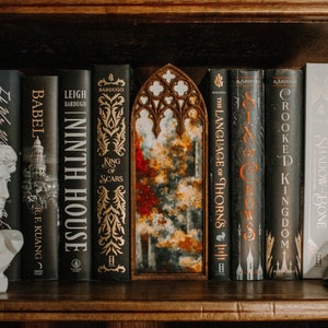 Op de afbeelding: Een boekenplank toont een verzameling boeken met donkere covers en gouden letters. Een witte buste van een klassieke figuur en een decoratief houten stuk met een gewelfde bovenkant zijn ook aanwezig. De boeken bevatten titels als "Ninth House" en "Six of Crows".