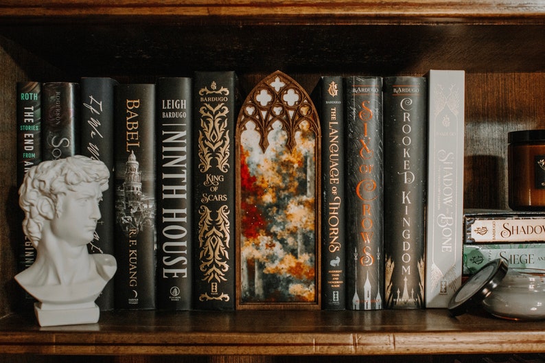Op de afbeelding: Een boekenplank toont een verzameling boeken met donkere covers en gouden letters. Een witte buste van een klassieke figuur en een decoratief houten stuk met een gewelfde bovenkant zijn ook aanwezig. De boeken bevatten titels als "Ninth House" en "Six of Crows".