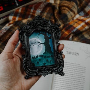 May include: A small, black framed picture with a spooky scene of a tree, full moon, and graveyard. The frame has ornate detailing. The word "Tavern" is visible on a book in the background. The image is held in a hand.