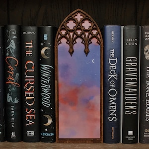 Op de afbeelding: Een boekenkast met een verscheidenheid aan boeken, waaronder "The Cursed Sea", "Wicked Deep", "Gravemaidens" en "The Deck of Omens". Een houten boekenlegger met een gegraveerde gotische boog en een geschilderd hemelscène bevindt zich in het midden.