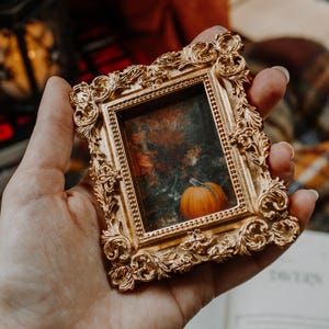 May include: A small, ornate gold picture frame with a miniature painting of a pumpkin. The frame has intricate scrollwork and a vintage aesthetic. The pumpkin is orange and sits in front of a fall-themed background.