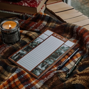 Peut inclure: Un bloc-notes à faire avec un design de scène de forêt en brun et blanc. Le bloc-notes est posé sur une couverture à carreaux avec une bougie allumée et deux stylos.
