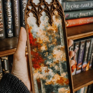 Op de afbeelding: Een houten spiegel met een gewelfde bovenkant, met een abstract landschap van oranje, rode en grijze tinten. De spiegel wordt voor een boekenplank vol boeken gehouden. Het frame heeft een sierlijk, gotisch design.
