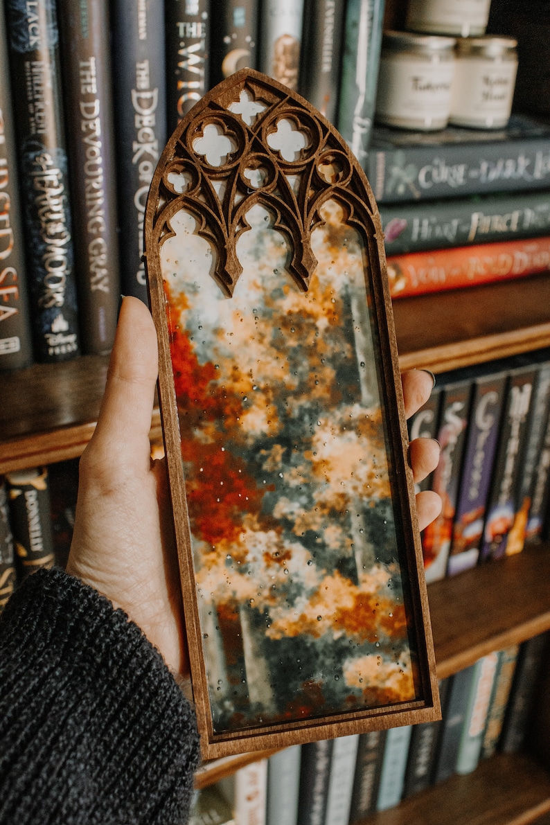 Op de afbeelding: Een houten spiegel met een gewelfde bovenkant, met een abstract landschap van oranje, rode en grijze tinten. De spiegel wordt voor een boekenplank vol boeken gehouden. Het frame heeft een sierlijk, gotisch design.