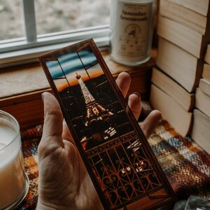 May include: A brown bookmark with a picture of the Eiffel Tower at night. The bookmark is held in a hand against a plaid background.