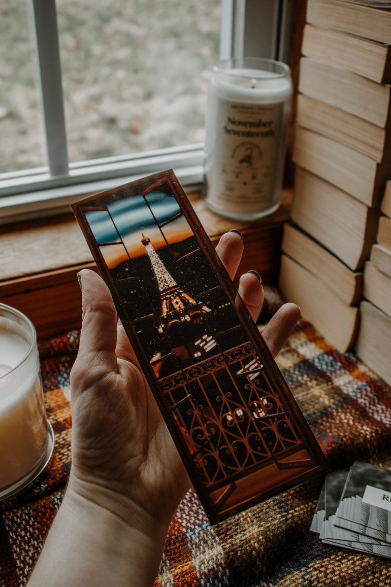 May include: A brown bookmark with a picture of the Eiffel Tower at night. The bookmark is held in a hand against a plaid background.
