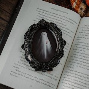 May include: A black framed oval picture featuring a ghost holding a candle, set against an open book. The ghost is white with two black eyes. The frame has an ornate, gothic design. The book pages are visible.